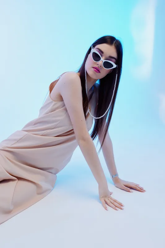 Young woman in a beige dress and white sunglasses poses on a light blue background. Concept of beauty, fashion, and modern style with a confident expression.