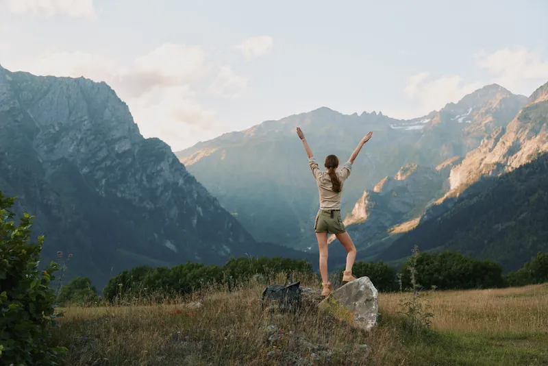 Young woman with long hair stands on a rock in a mountain valley raising her arms in joy during a sunny day, surrounded by tall peaks and open grassy fields.