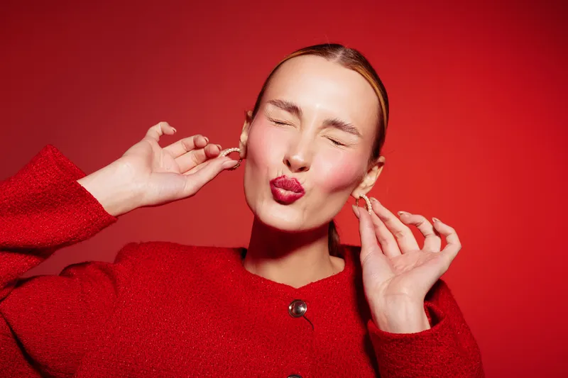 Young woman in red jacket makes playful face with closed eyes and puckered lips on red background. Concept of fun, fashion, and vibrant emotions in studio portrait.