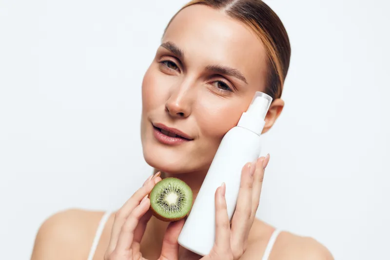 Caring woman with kiwi slice and bottle, skincare routine moment, clear skin, relaxed smile, soft light, natural colors, fresh mood, simple composition, hydration focus