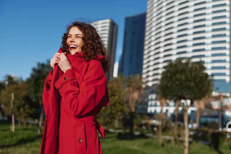 Smiling woman with curly hair wearing a red coat in an outdoor park. Urban skyline behind. Concept of fashion, lifestyle, and confidence. Bright daylight emphasizes warmth and happiness.
