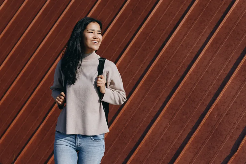 Female student with backpack walking beside a diagonal wooden wall, wearing a sweater and jeans, smiling and confident, capturing casual urban lifestyle in daylight with optimism and momentum