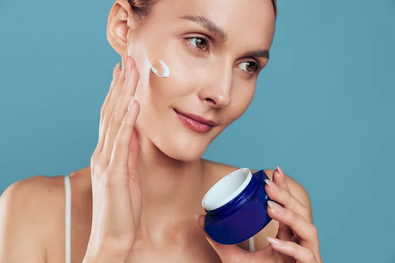 Woman applying face cream with fingers, holding a moisturizer jar, closeup on blue background, smiling in soft studio lighting. Skincare and beauty routine, self care and wellness.