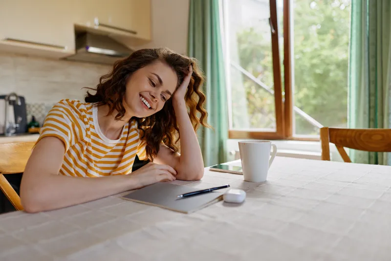 Bright indoor scene with a smiling woman at a home desk, writing in a notebook, warm daylight and a cozy kitchen background, relaxed lifestyle vibe