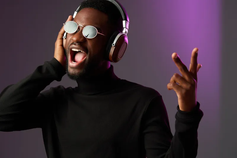 Excited young Black man in sunglasses and headphones enjoying music, expressing joy in a vibrant background with shades of purple