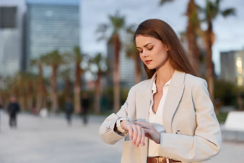 Business woman checking time on smartwatch outdoor with modern city background, wearing beige jacket, focused and professional emotion
