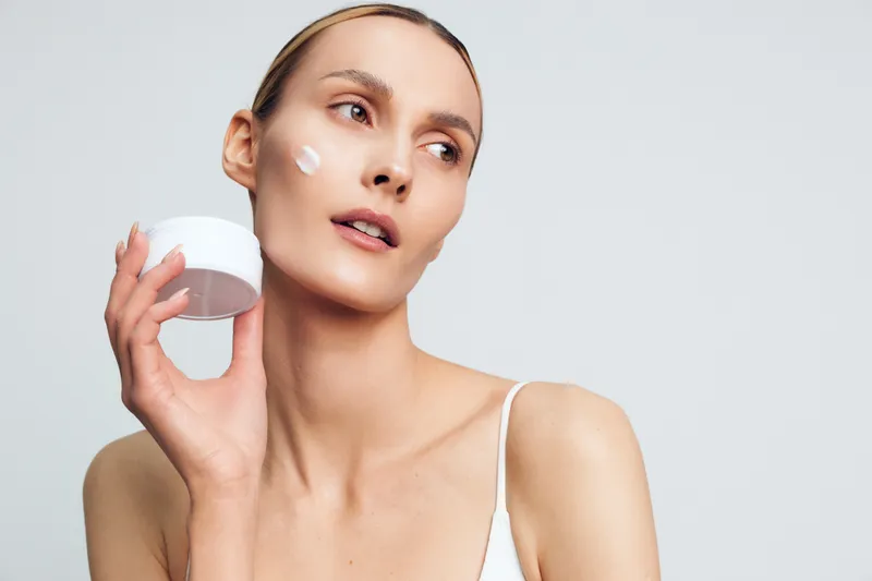 Young woman with clear skin applying moisturizer cream on face, skincare routine, natural beauty, clean background, healthy complexion, soft lighting, minimal makeup