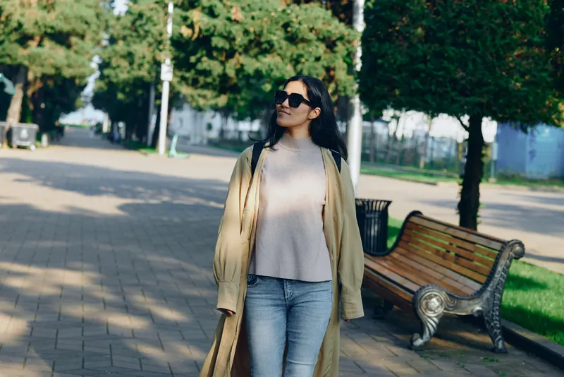 Woman fashion park stroll in casual coat and jeans under daylight with sunglasses, relaxed urban mood and candid street style in sunny park setting