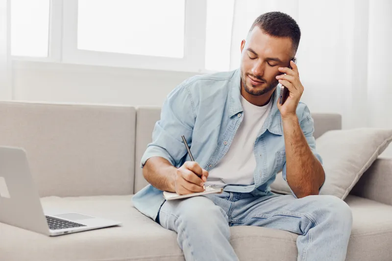 Thoughtful man talking on the phone while taking notes at home. Stylish, cozy setting with soft lighting, reflecting modern lifestyle and work-life balance.