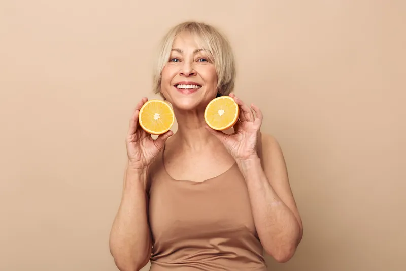 Smiling mature woman holds two orange halves near face on beige background. Concept of healthy lifestyle, natural beauty, vitamin C, and positive mood for senior wellness.
