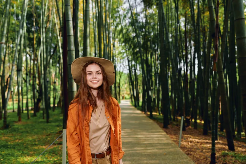 Woman smiling in straw hat and orange jacket walking on wooden path through bamboo forest, portrait of traveler in nature with sunlight filtering through tall bamboo stalks.