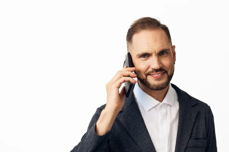 Professional business man on a plain colored background, speaking on a mobile phone, exuding confidence, focus, and calm energy with a poised stance and friendly expression.