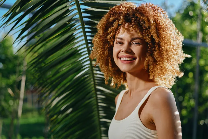 smiling woman with curly hair wearing white tank top outdoors near palm leaf in bright natural sunlight, healthy skin, happy expression, tropical background
