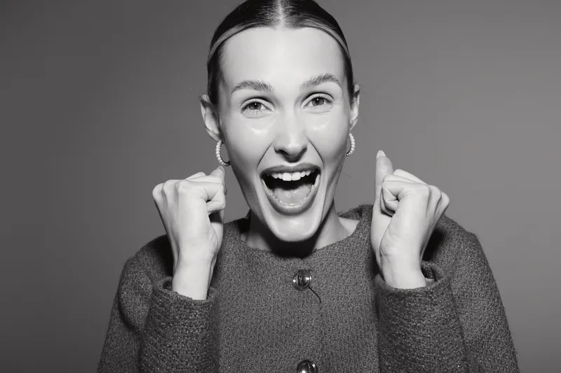 Excited Caucasian woman with light skin expresses joy with clenched fists and open mouth on grey background studio portrait.