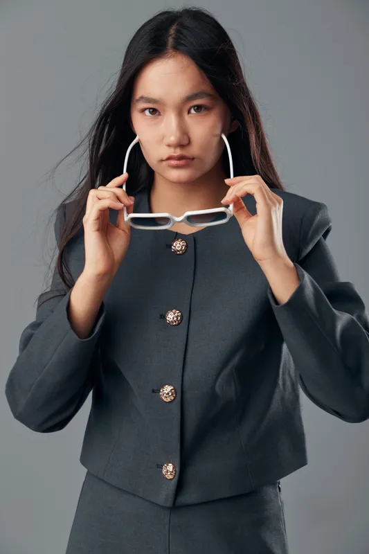 Woman wearing stylish dark suit holding eyeglasses with serious expression, standing on neutral background in studio for fashion or business portrait.