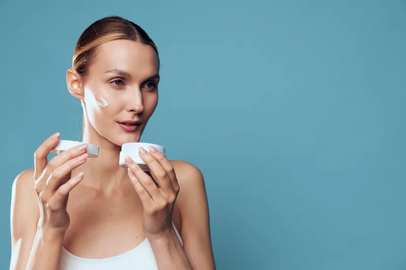 Young woman applies facial cream on clean skin holds jar with moisturizer looks focused isolated on blue background beauty skincare concept with natural makeup and casual hairstyle