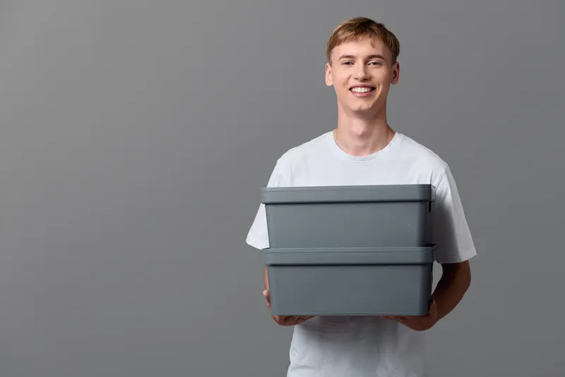 Delivery ready, a smiling young man holds two grey boxes for shipment, depicting packaging, storage and order processing in a clean studio setting for ecommerce display
