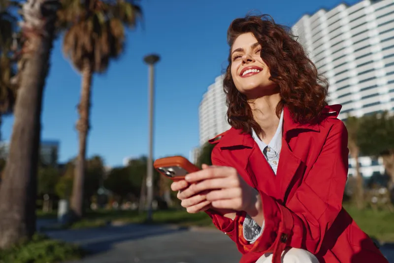 Young woman in red jacket smiling and holding smartphone outdoors on sunny day with palm trees and modern buildings in background.