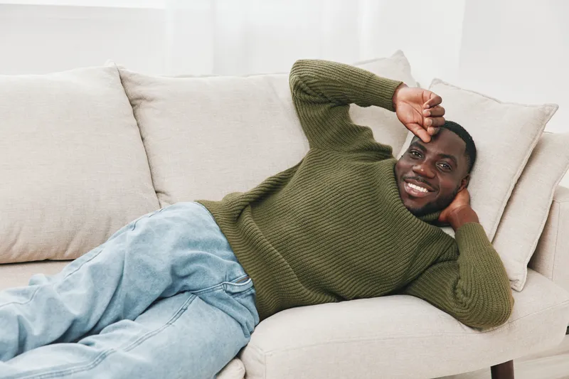 Relaxed young Black man in a cozy green sweater and denim lounging on a sofa, exuding happiness and comfort in a bright, airy living room