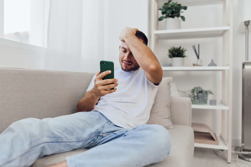 Young man sitting on a couch, looking at his phone with a concerned expression, surrounded by soft, modern decor. A relatable moment of digital stress in a cozy environment.