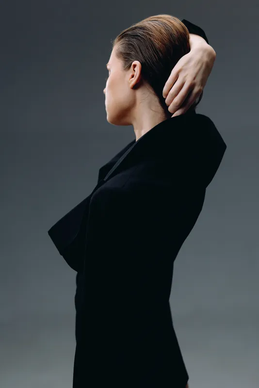 Woman in black outfit posing thoughtfully with hand behind head against gray background, elegant and minimalistic style