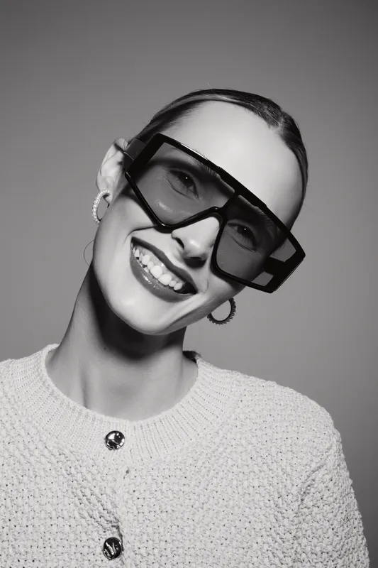 Smiling Caucasian woman with fair skin wearing large sunglasses and hoop earrings poses in knit sweater on gray background. Concept of fashion and style.