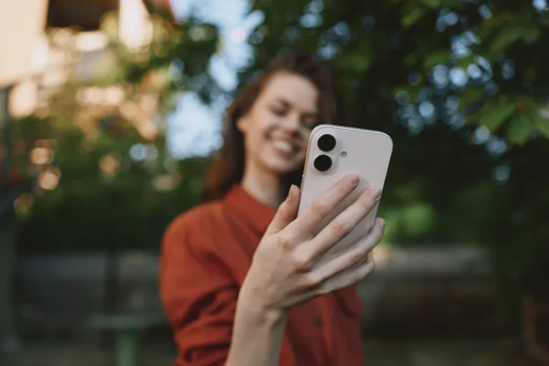 Outdoor selfie of a young woman smiling while taking a photo with a smartphone in a sunlit park with leafy background, casual rust top, relaxed pose