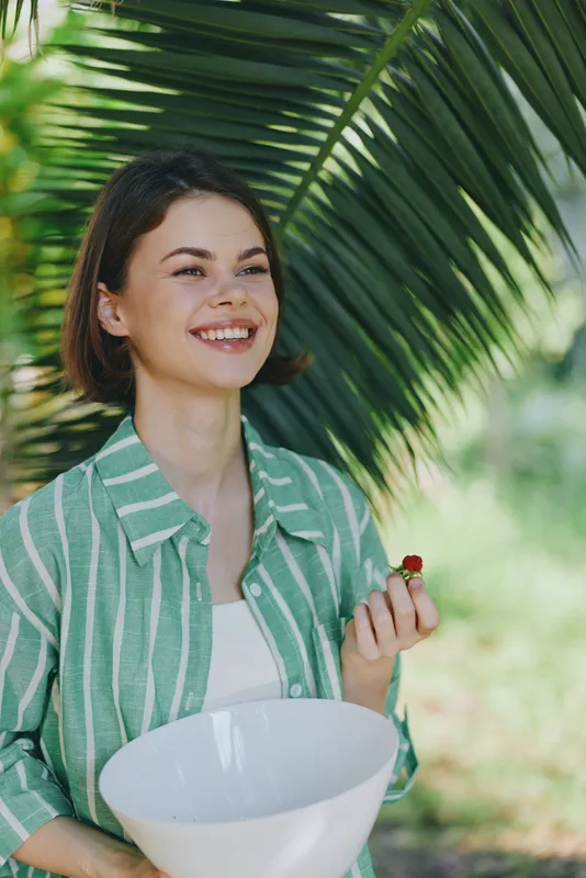 A cheerful woman stands outdoors among palm leaves, smiling warmly as she holds a white bowl, wearing a green striped shirt, radiating happiness and relaxed confidence in a sunny natural setting