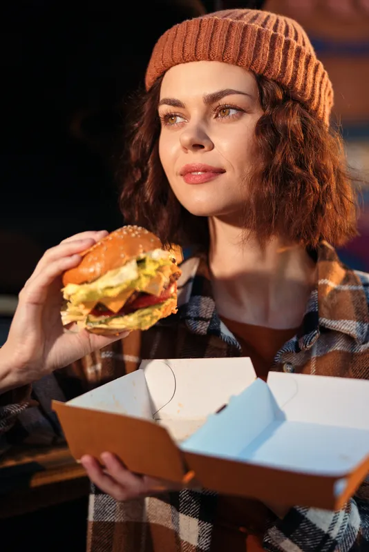 Woman portrait candid at golden hour holding burger and takeout box in urban street, beanie and plaid jacket, authenticity and candid lifestyle with golden hour glow and emotional storytelling.