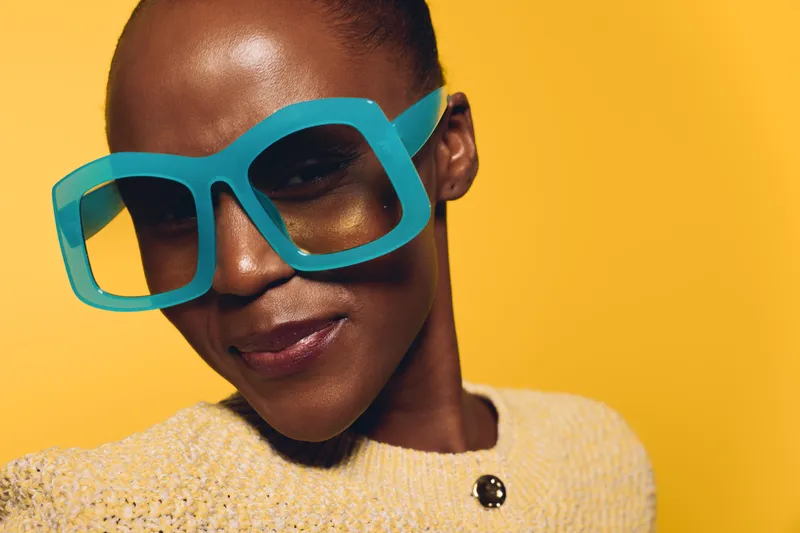 Close-up of a confident African woman with dark skin wearing oversized blue glasses and a beige sweater against a yellow background. Concept for fashion and style.