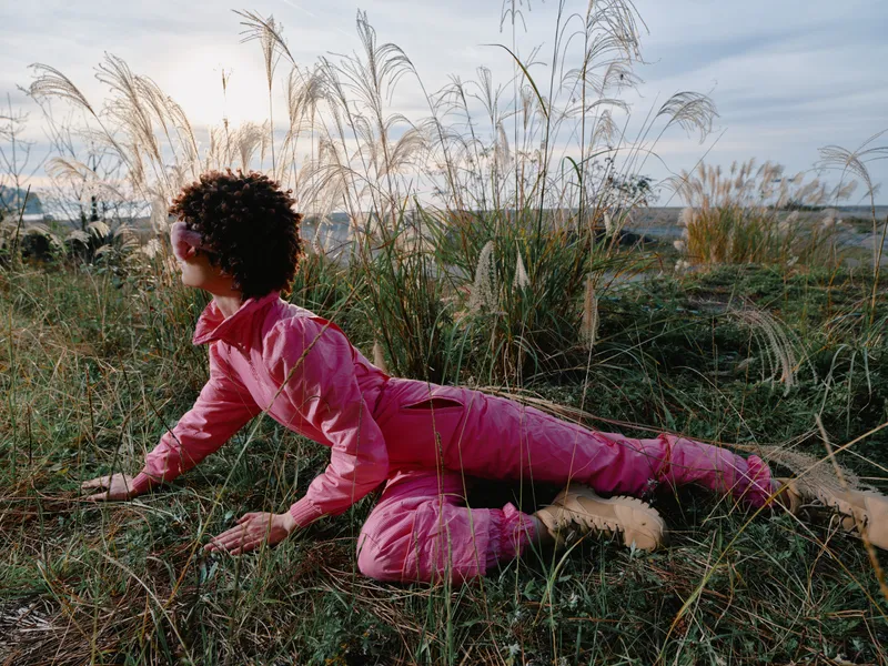Child in pink outfit crawls on a grassy field among tall wild grasses during golden hour, exploring nature with curiosity and gentle breeze, colorful earth tones around