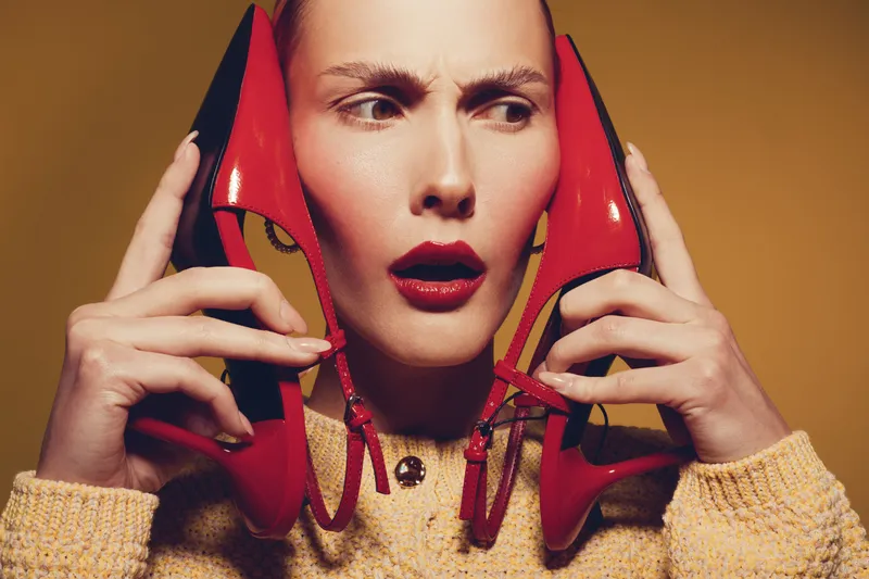 Confused woman with red lipstick holds red high heel shoes near her ears, wearing yellow sweater. Studio portrait with beige background. Concept of fashion, style, and unusual expression.