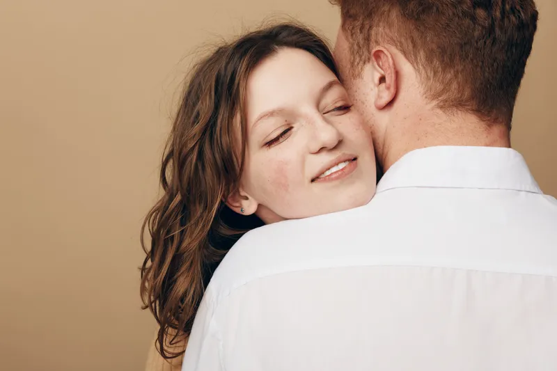 Young couple embraces tenderly, woman with closed eyes smiling softly while man faces away. Intimate moment of affection and love captured in warm neutral background.