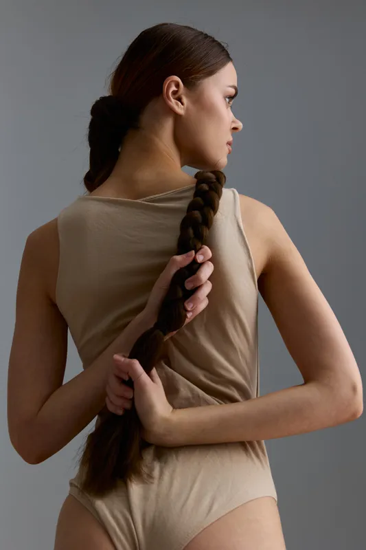 Young woman with long braided hair wearing beige bodysuit poses against grey background, showing elegant side profile and holding braid with both hands, minimalistic style.