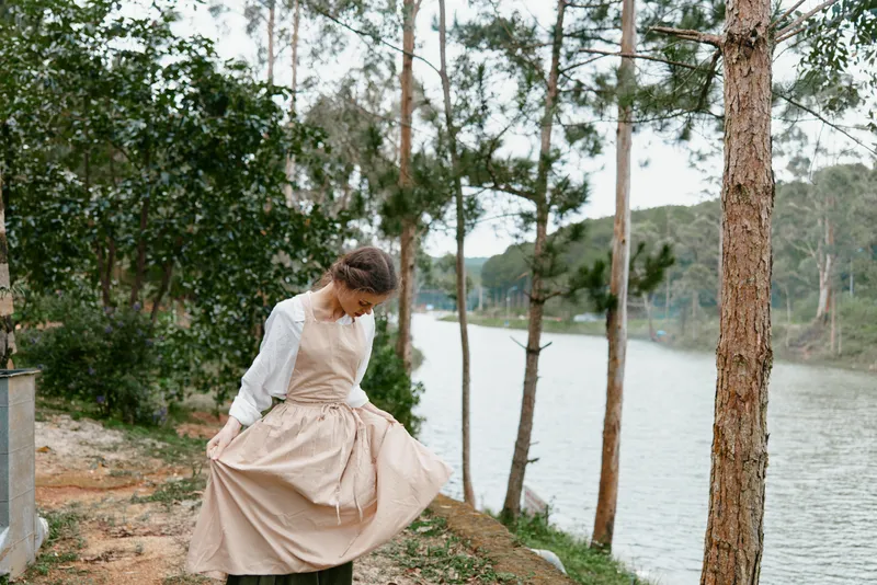 A young woman in a vintage dress stands by a serene lake, admiring her flowing skirt amid lush greenery, evoking a sense of tranquility and nature appreciation