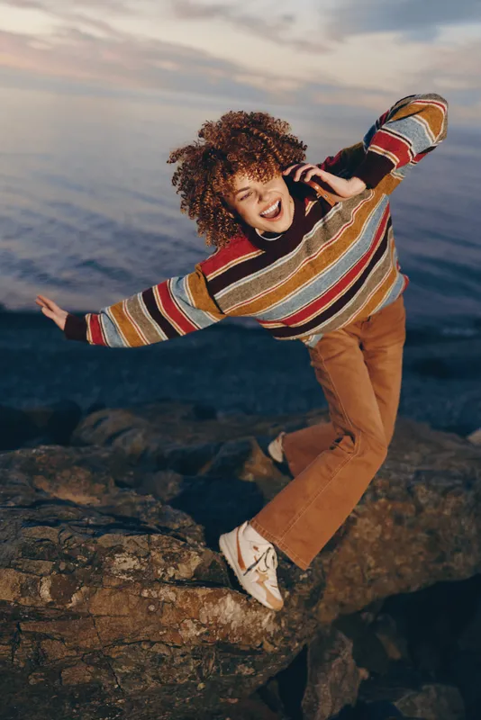 Woman lifestyle on rocky coast by ocean, laughing with wind in colorful sweater, relaxed stance at sunset, warm natural light, dramatic sky background