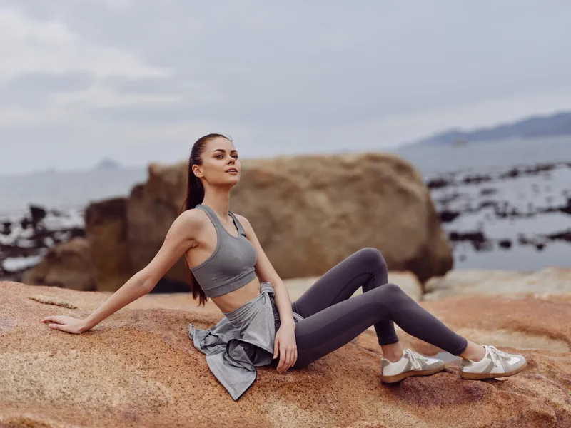 Young athletic woman in activewear sitting on a rock by the sea, showcasing health and fitness lifestyle, with a calm expression and soft natural lighting