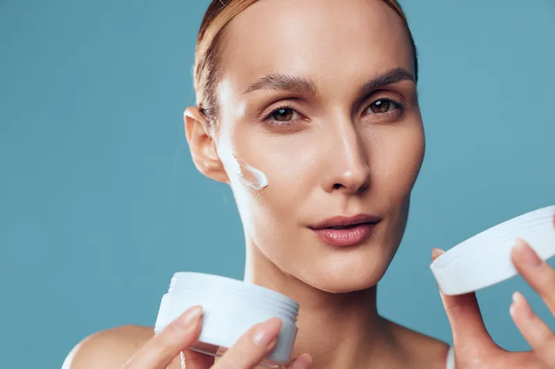 Woman skincare cosmetic beauty cream on face, holding white jar and applying moisturizer, closeup portrait on blue background, concept of hydration and selfcare for healthy skin
