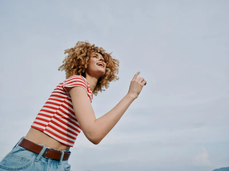Young girl with curly hair wears a red striped crop top and denim jeans, lifting an arm in a joyful, carefree moment beneath a clear blue sky.