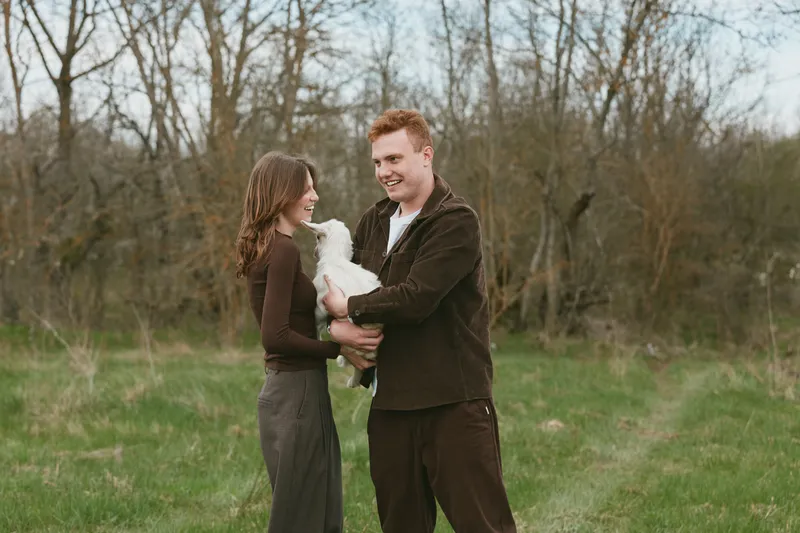 Smiling couple outdoors with a playful goat kid in a grassy field, held gently by the adults. Rustic countryside scene, warm natural lighting, celebrating family, nature, and farm life.