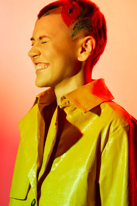 smiling woman with red braided hair wearing yellow textured coat against vibrant orange background, joyful expression, colorful portrait, warm lighting