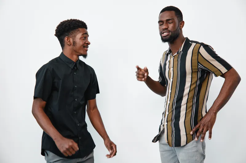 Two African American men with dark skin standing and talking together in casual clothes against white background. Young friends communicating and smiling indoors.