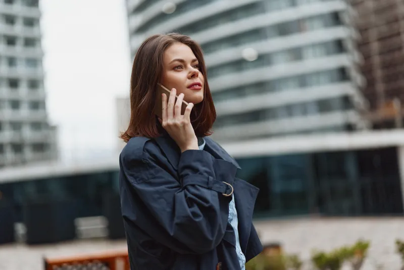 Young woman with shoulder length hair speaks on a smartphone in a modern urban courtyard, wearing a dark coat and casual outfit, under soft ambient light.