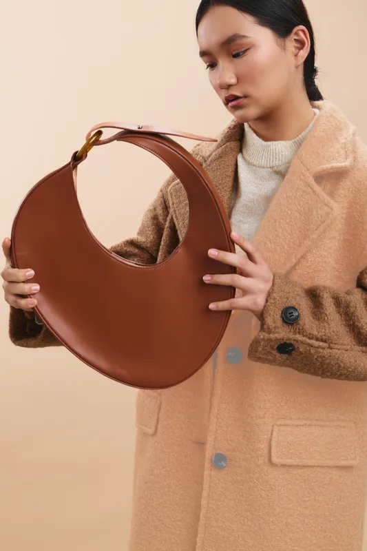 asian woman showcases a stylish handbag in a clean studio setting. she examines the curved leather bag with focus, conveying fashion and elegance against a neutral backdrop.