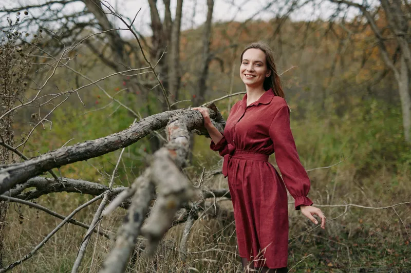 A candid portrait of a woman in a crimson dress leaning against a weathered fence, capturing authentic charm, natural light, and a serene autumn forest backdrop.