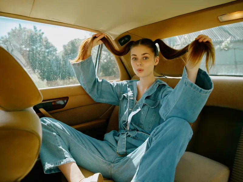 Young girl with long hair playfully posing in a car, wearing denim jumpsuit, reflecting joy and creativity in a sunny, natural setting.
