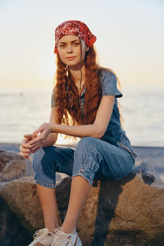 young woman in denim jumpsuit and red bandana sitting on rocks by sea during sunset, relaxed and thoughtful mood, casual style outfit