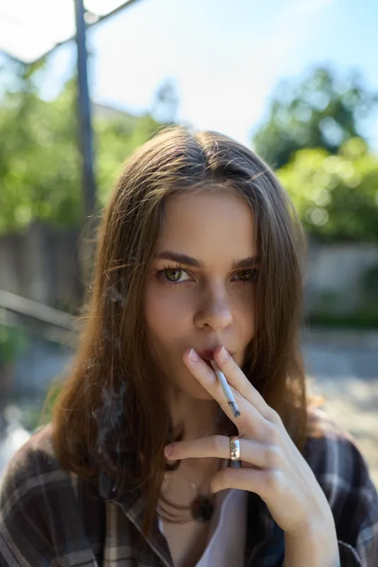 Smoking woman outdoors with a cigarette, a candid moment of calm concentration in casual attire, sunlit garden backdrop and relaxed gaze