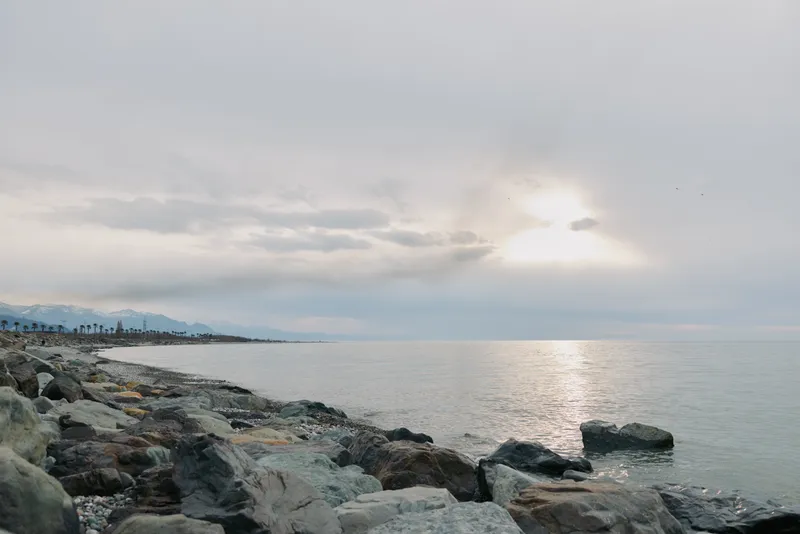 Rocky shoreline under cloudy sky at calm sea during sunset. Gentle waves touch stones with distant mountains and palm trees visible. Concept of nature, peace, and coastal landscape.