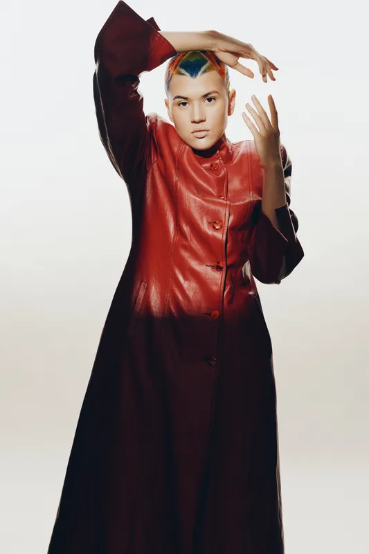 woman with colorful shaved hair in red leather coat posing with expressive hands against light background, modern fashion, striking style, bold look